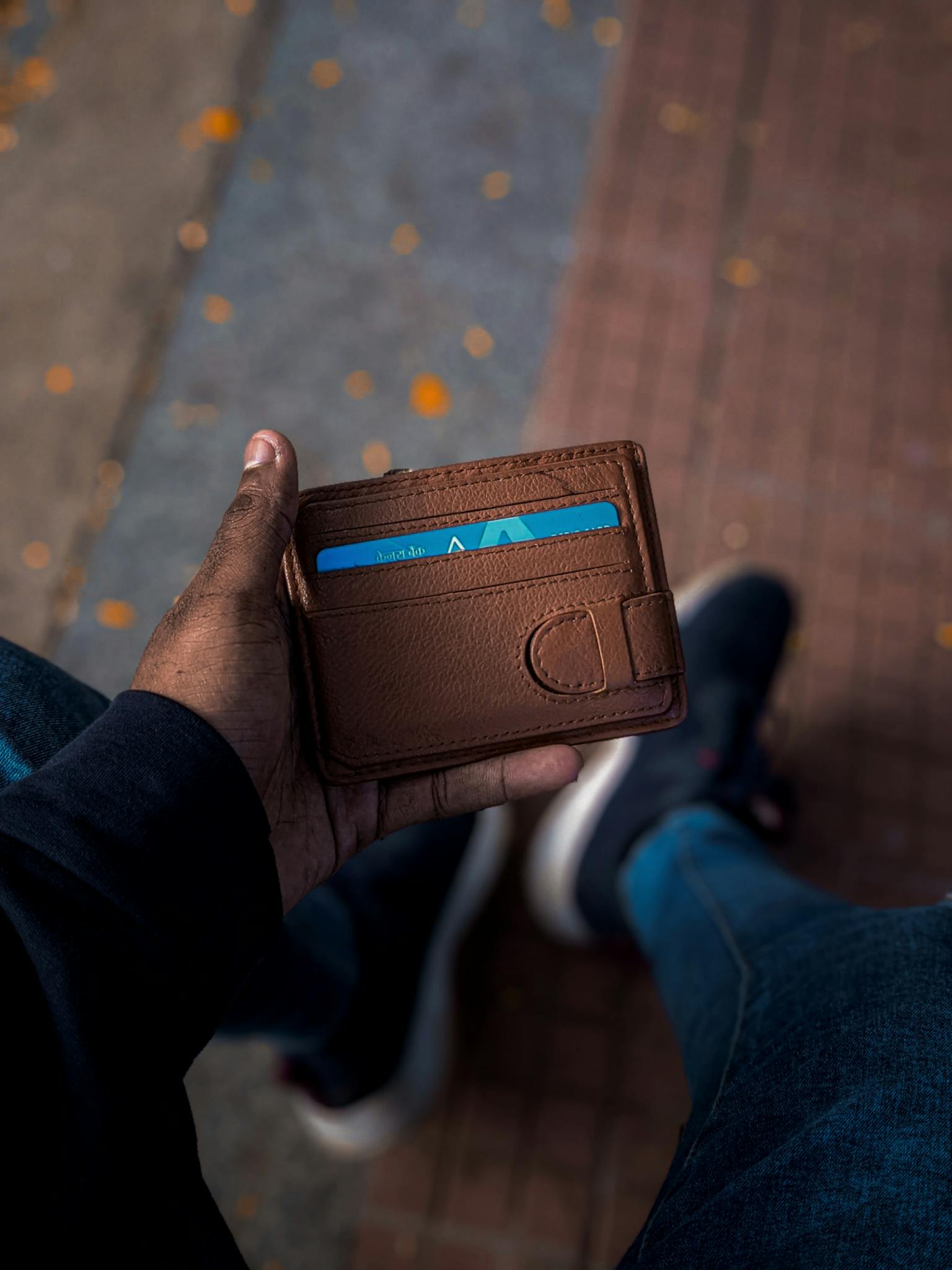 A close-up view of a man's hand holding a brown wallet with a credit card visible, captured on a sidewalk.